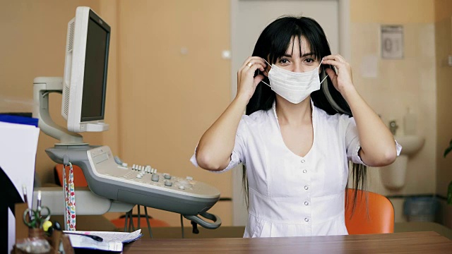 年轻的女医生微笑着看着摄像机，戴上了一个保护性的外科口罩。她正坐在医生办公室的桌旁。在4 k拍摄视频下载