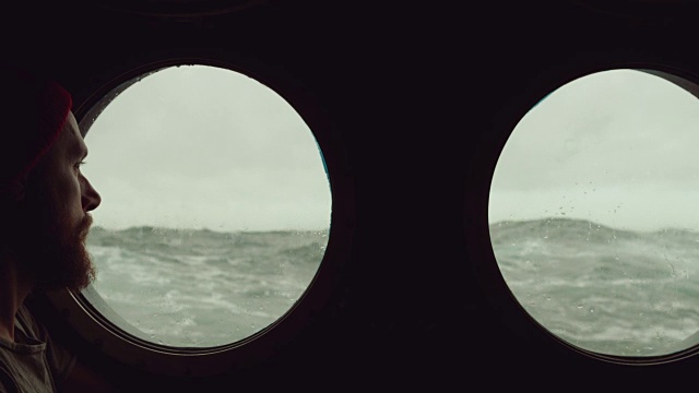 在波涛汹涌的海面上，一名长胡子的水手站在一艘船的舷窗前视频素材