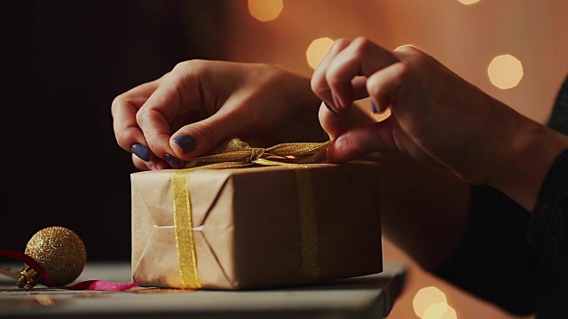 特写的背景圣诞灯妇女的手系在牛皮纸礼物上的丝带。背景是模糊的散景视频下载
