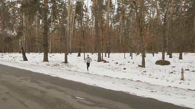 一个留着胡子的年轻运动员沿着森林附近的冬季道路奔跑视频素材