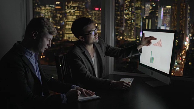 两个在办公室工作到很晚的商人。与电脑显示器讨论利润。图表，图表和城市灯光背景。视频素材