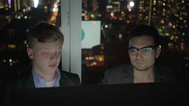 两个在办公室工作到很晚的商人。与电脑显示器讨论利润。图表，图表和城市灯光背景。视频素材