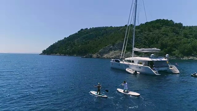 空中跟踪拍摄的一个两个女孩站着冲浪板附近的帆船。豪华白色游艇停泊在美丽的绿色岩石海岸。视频素材