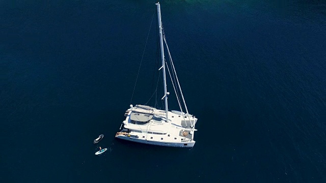 空中从上往下拍摄的一艘白色大帆船抛锚在一个海湾，旁边有一个男人站着玩滑板。海洋是深蓝色和美丽的。视频下载
