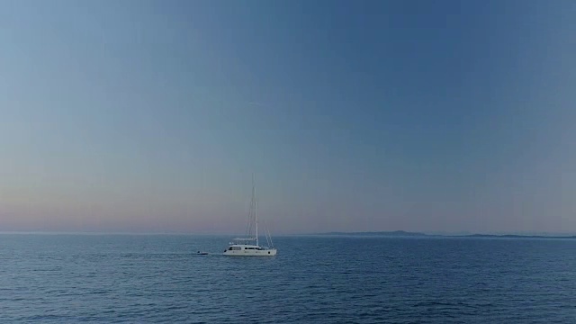 航空延时弧线拍摄的白色帆船旅行在一个平静的海上与海岸边的山丘可见。早上/晚上好。视频素材