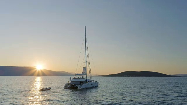 航拍一艘航行双体游艇在平静的海面上旅行，太阳从山后升起。视频素材