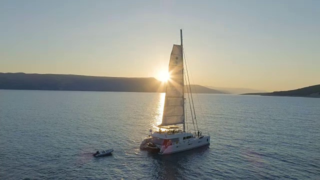 航拍的帆船双体游艇与升起的帆通过平静的海洋与太阳升起后的山。人们在甲板上休息。视频素材