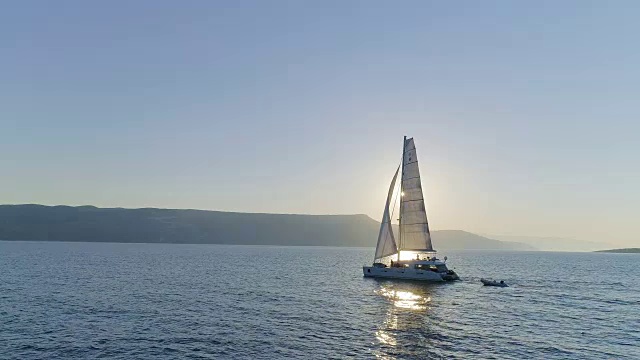 航拍长镜头一艘帆船双体游艇与升起的帆通过平静的海洋与太阳升起与海岸山丘可见。人们在甲板上休息。视频素材
