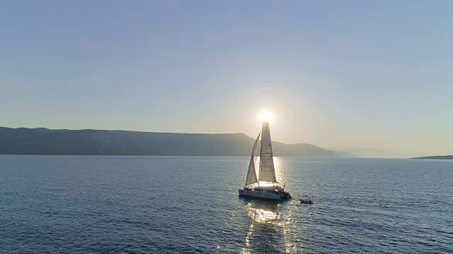 航拍长镜头一艘航行的双体游艇与升起的帆旅行通过公海。阳光照耀着海岸边的山丘。视频素材