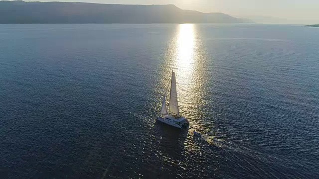 鸟瞰图的帆船双体游艇与提升的帆旅行通过公海。阳光照耀着海岸边的山丘。视频素材