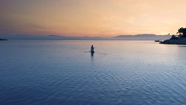 航拍的一个美丽的女人与一个站立板。女人的剪影与粉红色的日落和海岸山可见视频素材