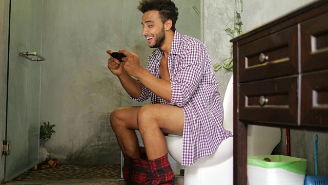 坐在马桶上的男人用手机用智能手机年轻的拉丁男人在玩游戏视频素材