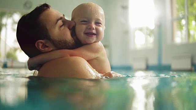 可爱的小婴儿和他的爸爸在游泳池里上游泳课。父亲把儿子抱在手里，拥抱着他。小男孩开心地笑着视频素材
