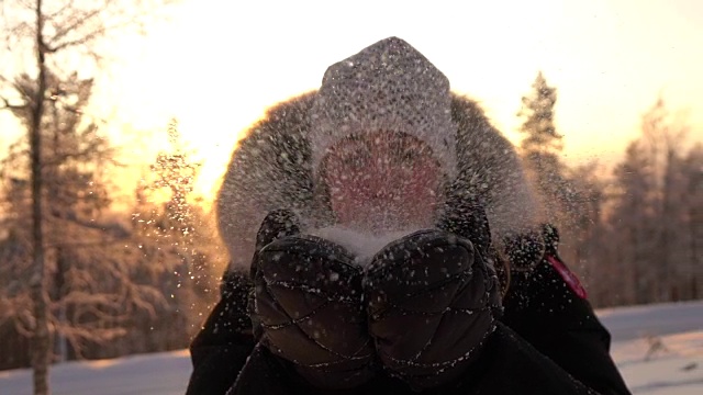 慢镜头:在神奇的冬天，年轻女子在夕阳下吹着雪花视频素材
