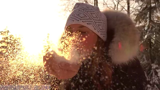 慢镜头:一个年轻女子在神奇的冬日夕阳下吹着闪闪发光的雪花视频素材