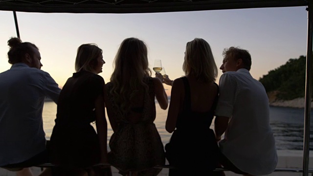 五名年轻人在游艇上碰杯和欣赏海景日落的后视图。视频素材