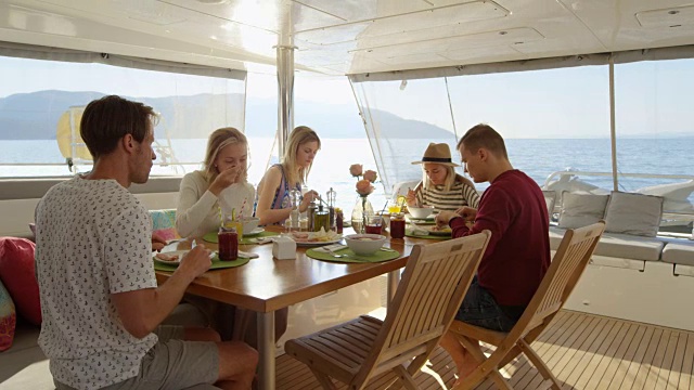 一群年轻漂亮的人在游艇上吃健康早餐，他们在友好地聊天。他们有各种各样的菜和冰沙上桌。海是背景。视频下载