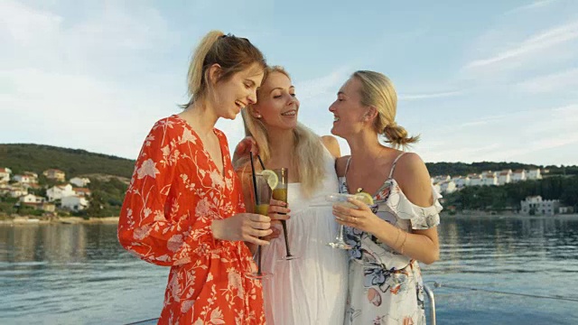 三个年轻漂亮的女人穿着夏装在游艇上喝鸡尾酒，欢笑，享受美好时光。背景是平静的大海和小岛村庄。视频素材