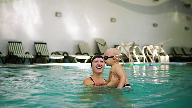 年轻的母亲抱着戴着防护眼镜的小男孩在游泳池里教他如何游泳。宝宝用脚在水里踢，开始在室内游泳池游泳视频素材