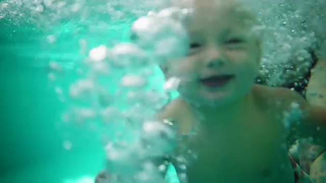 可爱的金发小孩在妈妈抱着他的时候潜入水中。他妈妈正在教他游泳。一个水下拍摄视频素材