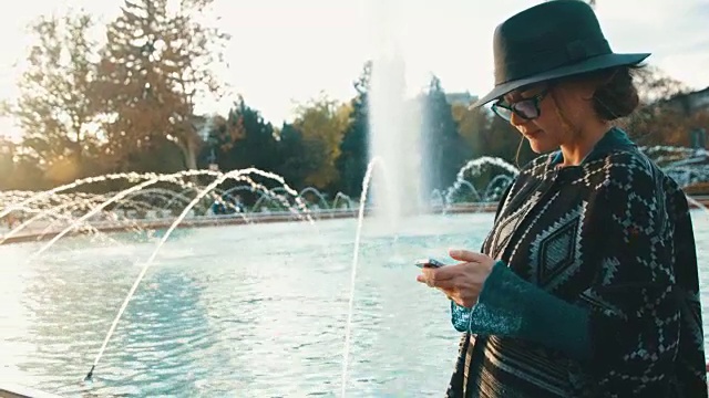 美丽的女孩站在喷泉前，用她的智能手机视频下载