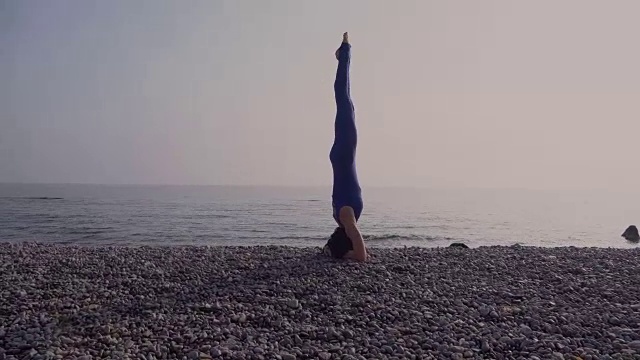 年轻女子在海滩上练习瑜伽在海上在惊人的日出。健身、运动、瑜伽和健康生活理念。女孩做着倒立和麻绳视频下载