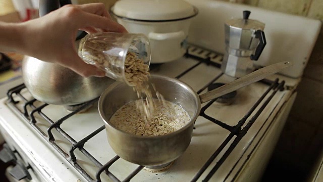 健康的素食早餐，燕麦粥。一个白人妇女的手把大的燕麦片从玻璃倒进煤气炉上的金属锅里。有机早餐，强健肌肉视频素材