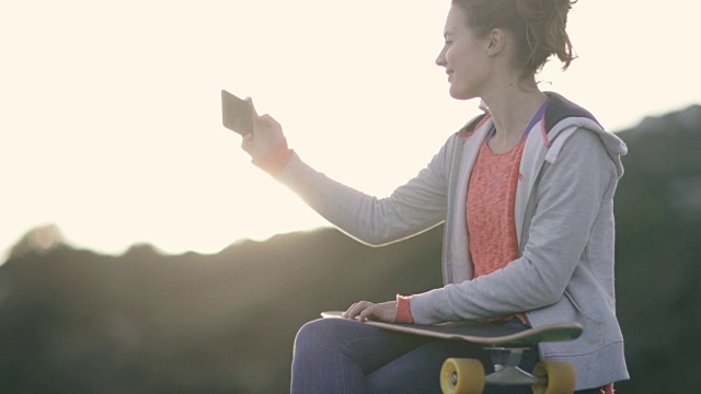 年轻的成年女性滑板运动员在日落时给朋友发短信视频素材