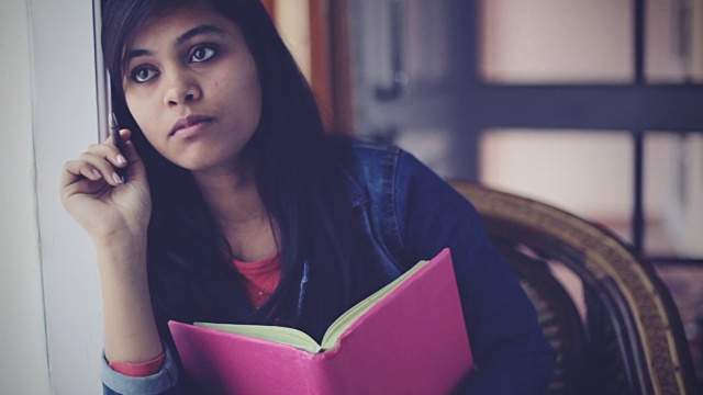安详的亚洲年轻成年学生靠近窗户，拿着书。视频素材