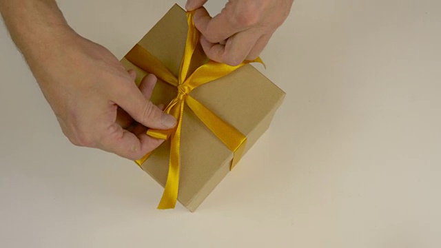 快速运动时间推移。包装礼盒。白种人手工包装礼盒。男人的手在一个棕色的硬纸盒上系上一条丝带。俯视图特写。金色的丝带，黄色的带子。视频素材
