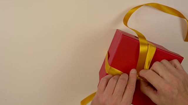 快速运动时间推移。包装红色礼品盒。白种人手工包装礼盒。男人的手在一个红色的硬纸盒上系上一条丝带。俯视图特写。金色的丝带，黄色的带子。视频素材