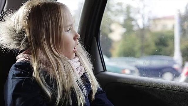 一个金发小女孩坐在车后座上。悲伤的孩子看着车窗，打哈欠。视频素材