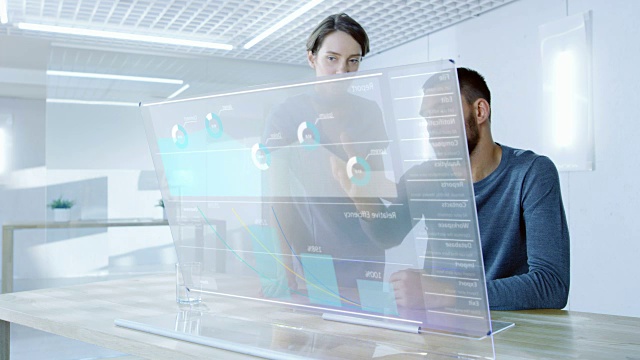 在不久的将来，男性和女性办公室工作人员讨论在透明电脑显示器上显示的图形统计。漂亮的人说话。屏幕上的交互式图表。视频素材