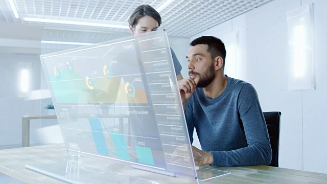 在不久的将来，男性和女性办公室工作人员讨论在透明电脑显示器上显示的图形统计。漂亮的人说话。屏幕上的交互式图表。视频购买