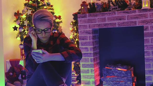 一名年轻女子盘腿坐在家中圣诞树前的地板上查看手机短信。视频素材