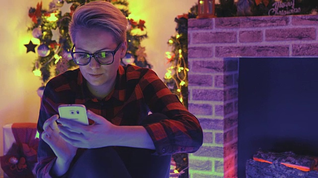 一名年轻女子盘腿坐在家中圣诞树前的地板上查看手机短信。视频素材
