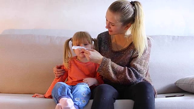 年轻的母亲和女儿坐在沙发上，擦拭婴儿的鼻子视频下载