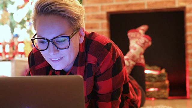 圣诞节期间，一位年轻女子在家里放松，她在客厅圣诞树前的地板上用笔记本电脑上网。视频下载