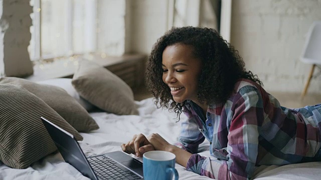 卷毛非洲裔美国女孩躺在家里的床上用笔记本电脑分享社交媒体视频素材