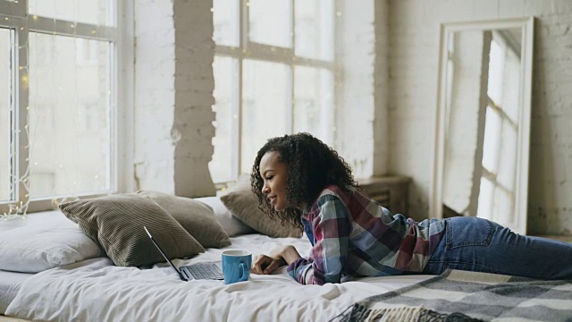 迷人的混血少女躺在家里的床上用笔记本电脑分享社交媒体大笑视频素材