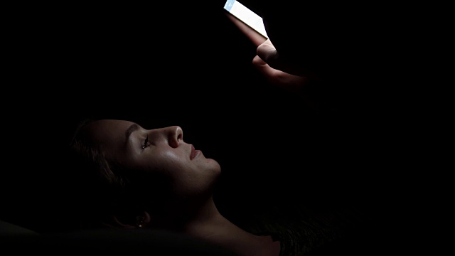 一个女人躺在沙发上，在黑暗的房间里使用她的手机，在黑暗中与智能手机聊天，与朋友在线交流的特写镜头视频下载