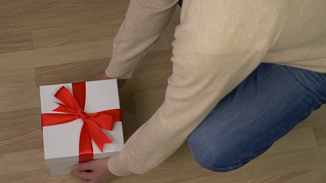 男子带来白色礼盒与红色丝带蝴蝶结，并把它放在地板上。木地板在室内。俯视图高角度。穿牛仔裤的男人拿着礼品盒走了。视频素材