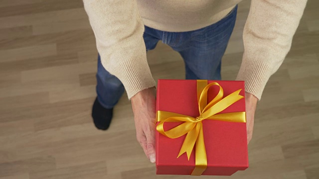 一名身穿米色毛衣的白人男子手里拿着一个礼品盒。俯视图的男人在休闲米色毛衣。人类给了礼物。锁定。红色的礼盒。高角度。铜。视频素材