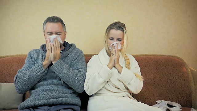 慢镜头:一个男孩和一个女孩坐在家里的沙发上打喷嚏，用手帕擦鼻涕。视频素材