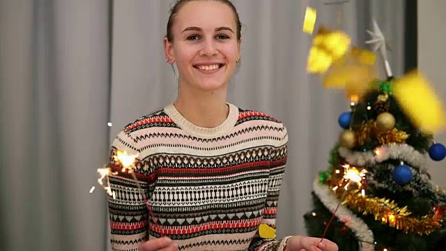 金色的五彩纸屑落在一位微笑着的年轻女子身上，她的背景是圣诞树和花环，她拿着烟花看着镜头。Slowmotion拍摄视频素材
