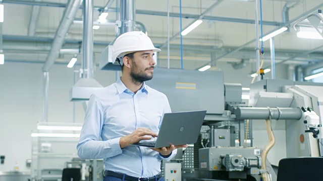 戴安全帽的总工程师手拿笔记本电脑穿过轻型现代工厂。现代工业环境下的成功英俊男人。视频素材