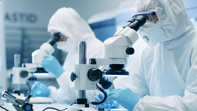 两名工程师/科学家/技术员穿着无菌洁净服使用显微镜进行组件调整和研究。他们在一家电子元件制造厂工作。视频素材
