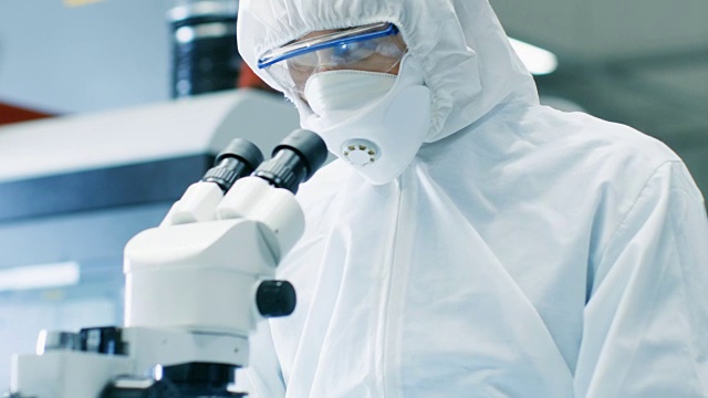 工程师/技术员/科学家在显微镜下检查组件的特写。他在一个项目中穿着无菌工作服和防护眼镜。视频素材