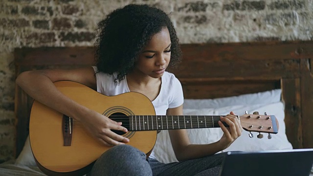 年轻的卷曲的混血女孩集中学习弹吉他使用笔记本电脑坐在家里的床上视频素材