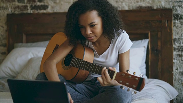 迷人的非洲裔美国少女集中学习弹吉他使用笔记本电脑坐在家里的床上视频素材